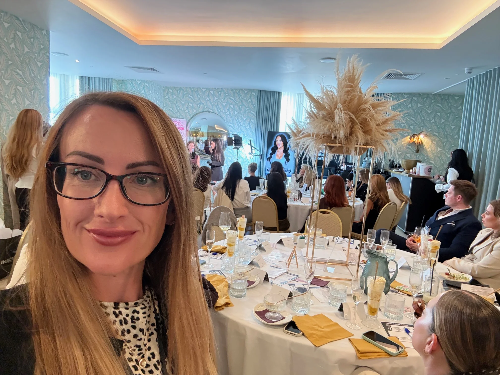 Alexis Drake at an aesthetics industry event, with attendees seated around tables and a presentation in the background